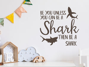 Wall decal for kids in a brown color that says ‘Always Be You Unless You Can Be A Shark’ on a kid’s room wall. 