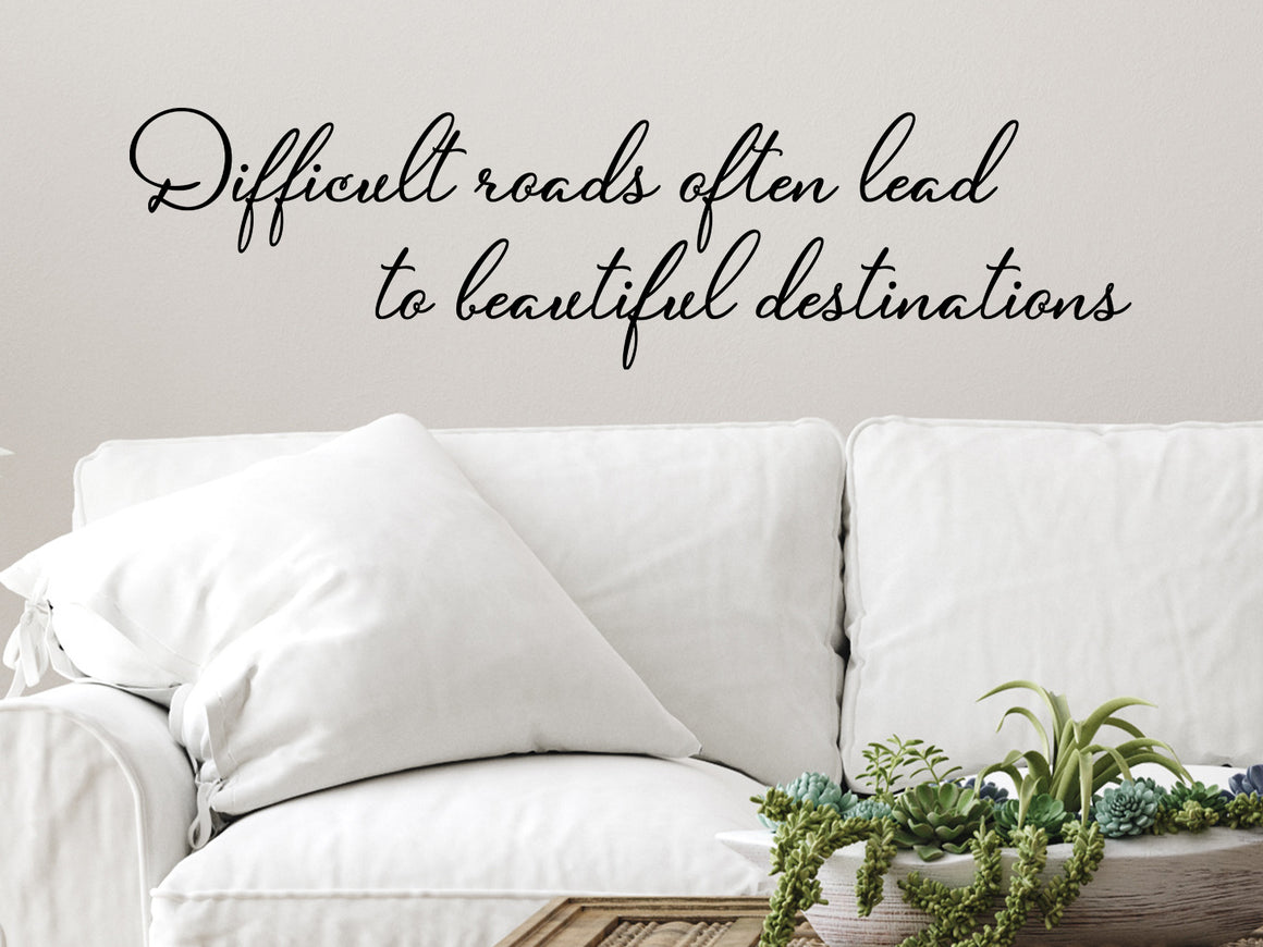 Living room wall decals that say ‘Difficult Roads Often Lead To Beautiful Destinations’ in a cursive font on a living room wall. 