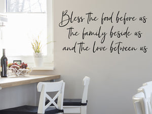 Wall decals for kitchen that say ‘Bless The Food Before Us The Family Beside Us And The Love Between Us’ in a script font on a kitchen wall.