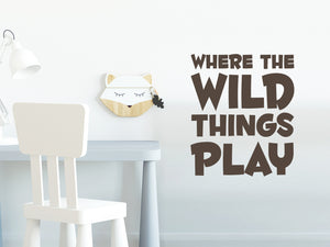 Wall decal for kids in a brown color that says ‘Where The Wild Things Play’ in a bold font on a kid’s room wall. 
