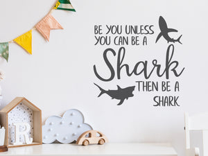 Wall decal for kids in a grey color that says ‘Always Be You Unless You Can Be A Shark’ on a kid’s room wall. 