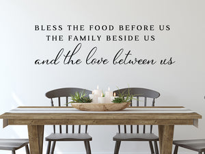 Wall decals for kitchen that say ‘Bless The Food Before Us The Family Beside Us And The Love Between Us’ in a print font on a kitchen wall.