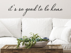 Living room wall decals that say ‘It's So Good To Be Home’ in a cursive font on a living room wall. 