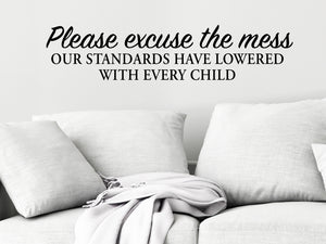 Living room wall decals that say ‘Please Excuse The Mess Our Standards Have Lowered With Every Child’ in a script font on a living room wall. 